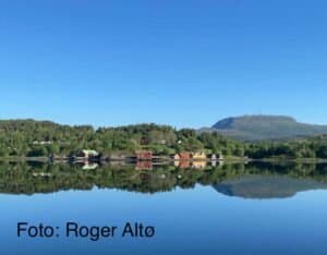 En rolig innsjø reflekterer grønne åser, spredte hus og et fjernt fjell under en klar blå himmel.