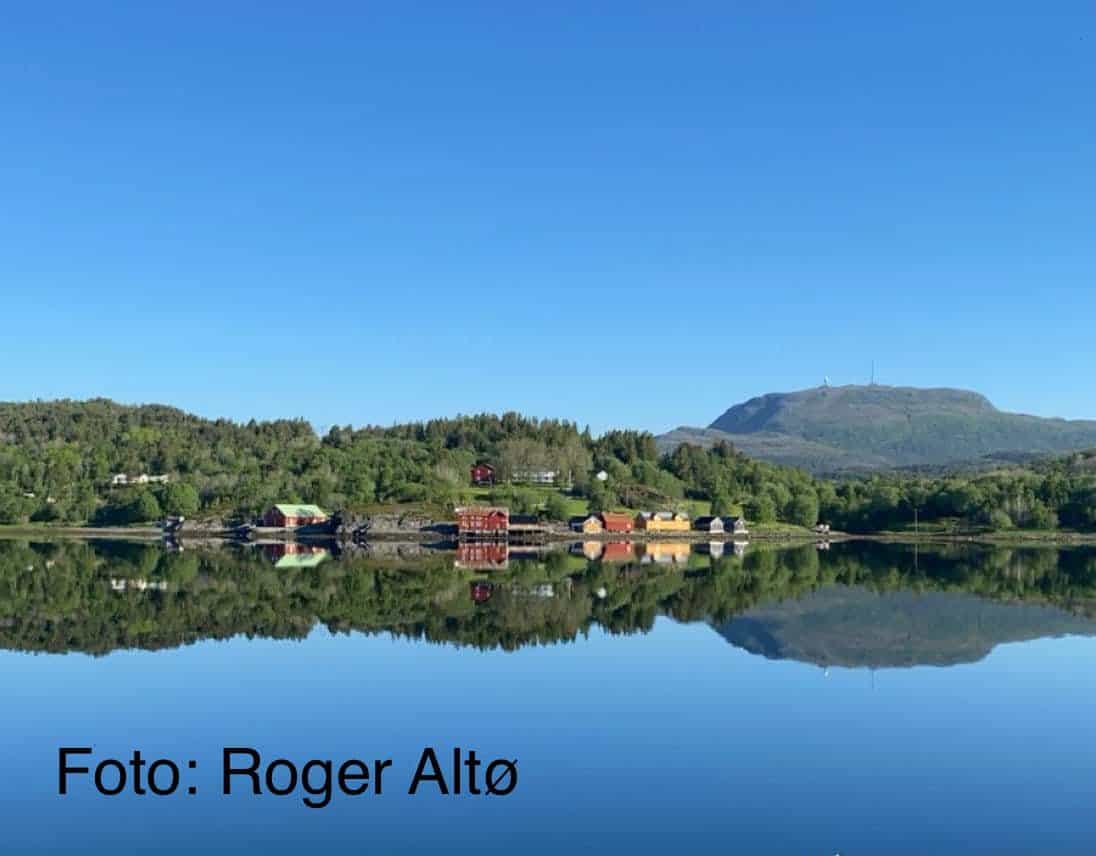 En rolig innsjø reflekterer grønne åser, spredte hus og et fjernt fjell under en klar blå himmel.