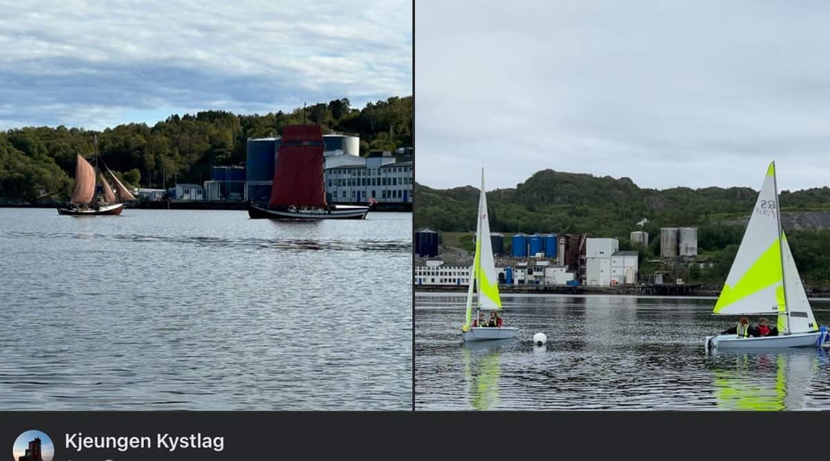 To seilbåter med mørkerøde seil og to små seilbåter med hvite og gule seil ligger på stille vann i nærheten av en industrikant og trekledde åser.